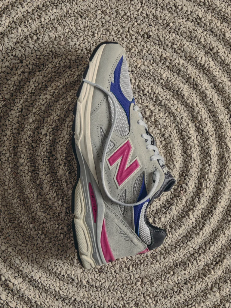 Gray New Balance sneaker with pink and blue accents lying on beige gravel, viewed from above