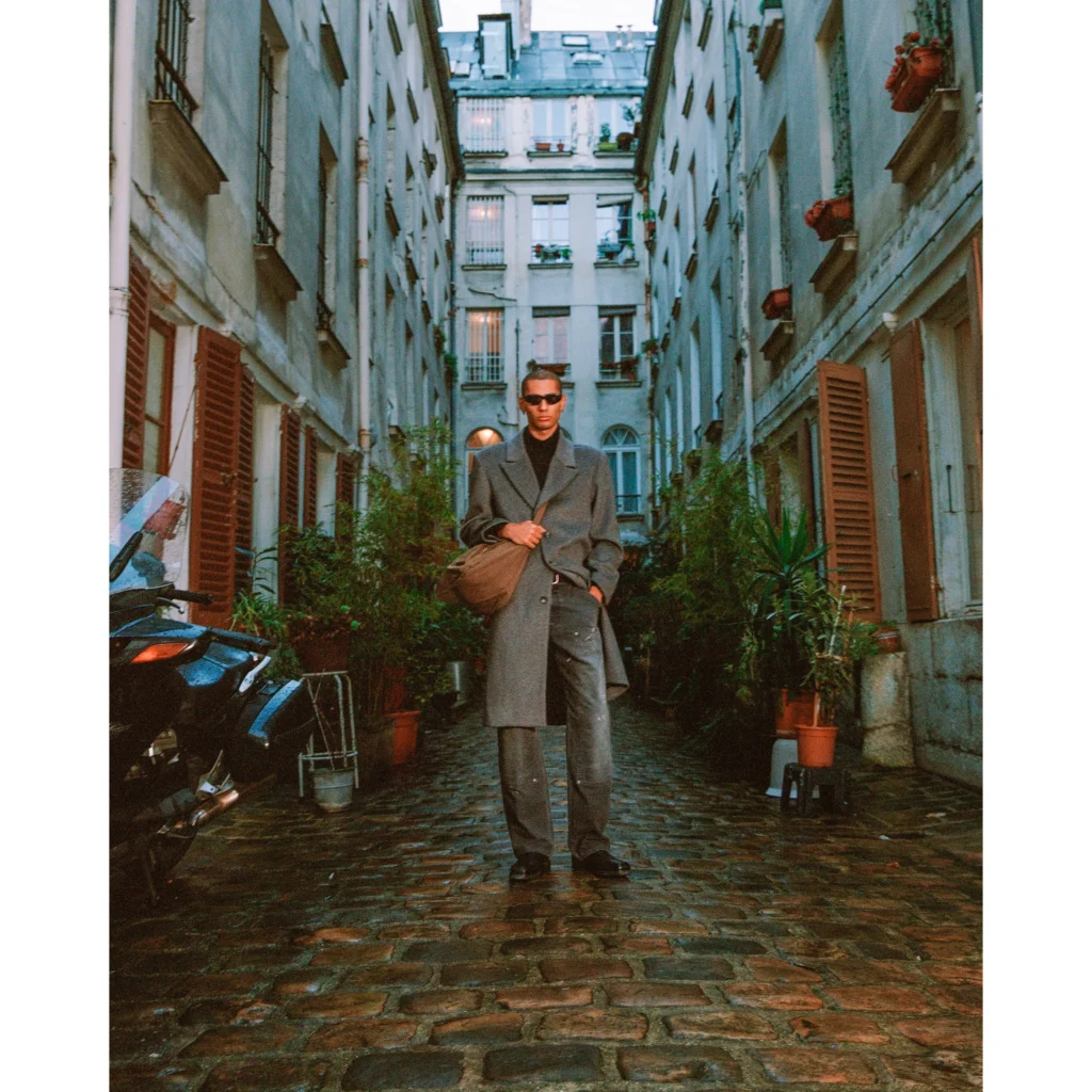 Man in a gray overcoat walks through a narrow cobblestone courtyard lined with potted plants and tall buildings