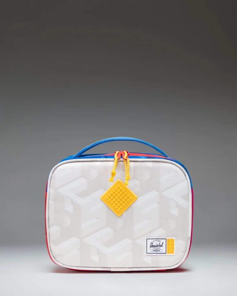 White patterned Herschel lunch bag with blue handle, red trim, yellow zipper pulls and hanging tag on gray background