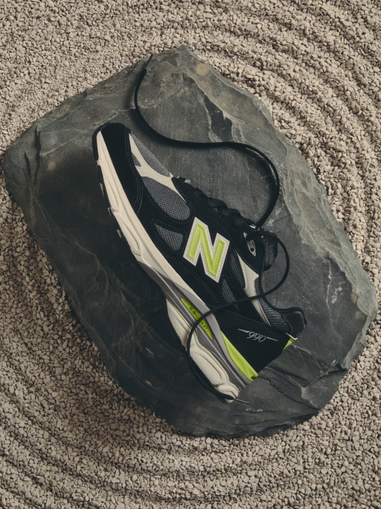 Black and white New Balance sneaker with neon green accents and loose laces on a dark rock surrounded by pale gravel