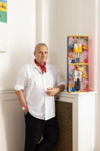 Man in a white shirt and red neckerchief stands beside boxed Barbie and Ken dolls in a bright room