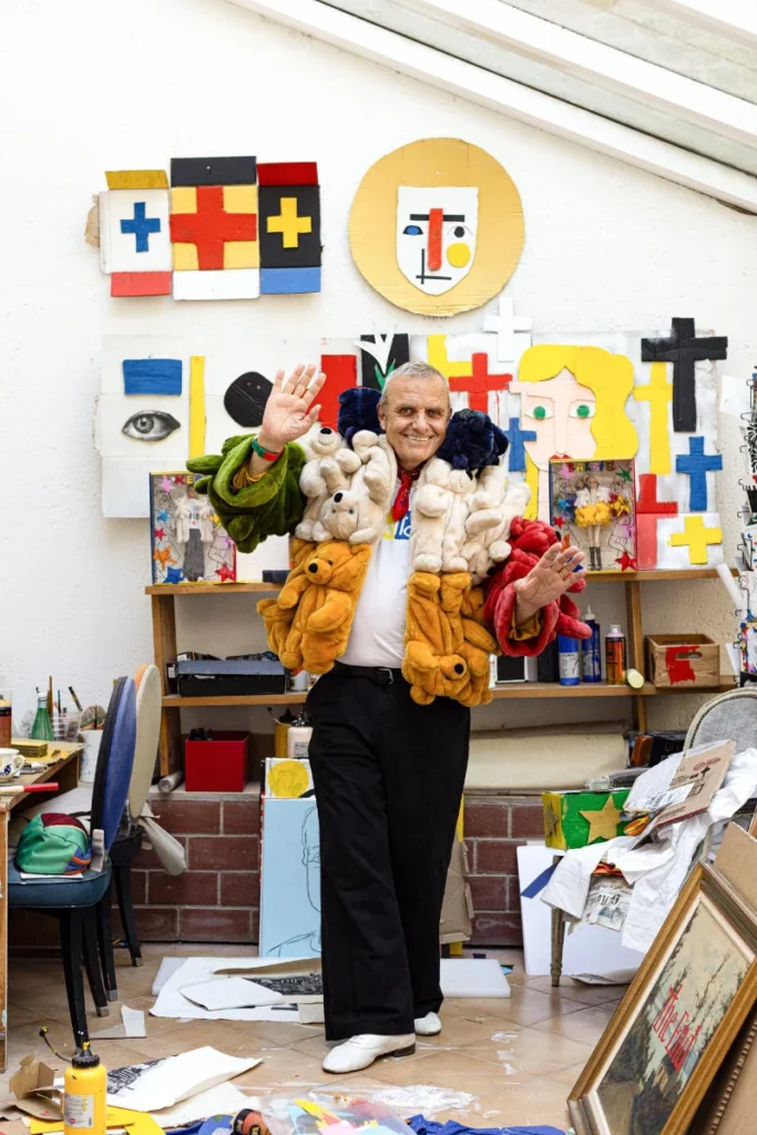Man wearing a multicolored teddy bear coat waves in an art-filled studio
