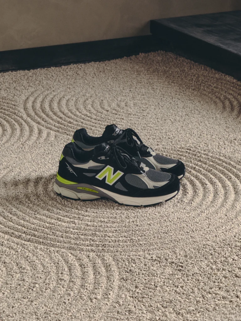 Pair of black New Balance sneakers with gray panels and neon green accents on a textured rug beside a dark platform bed