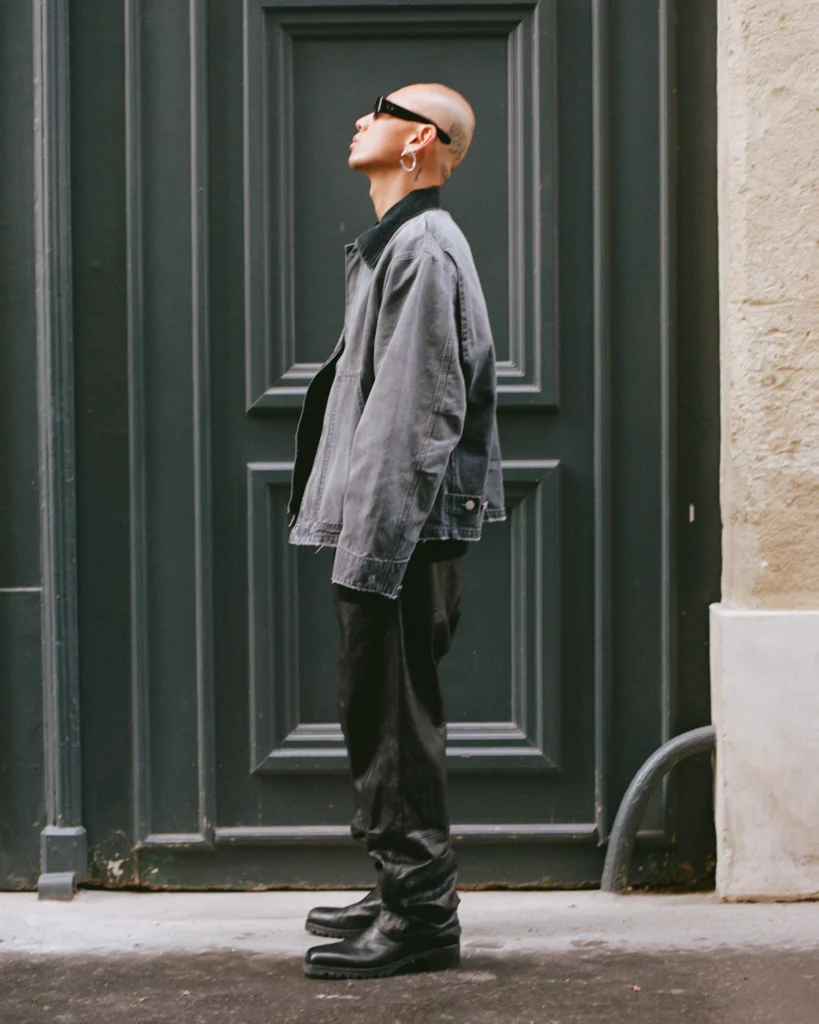 Bald person in sunglasses, gray jacket, black pants and boots standing before a dark paneled door