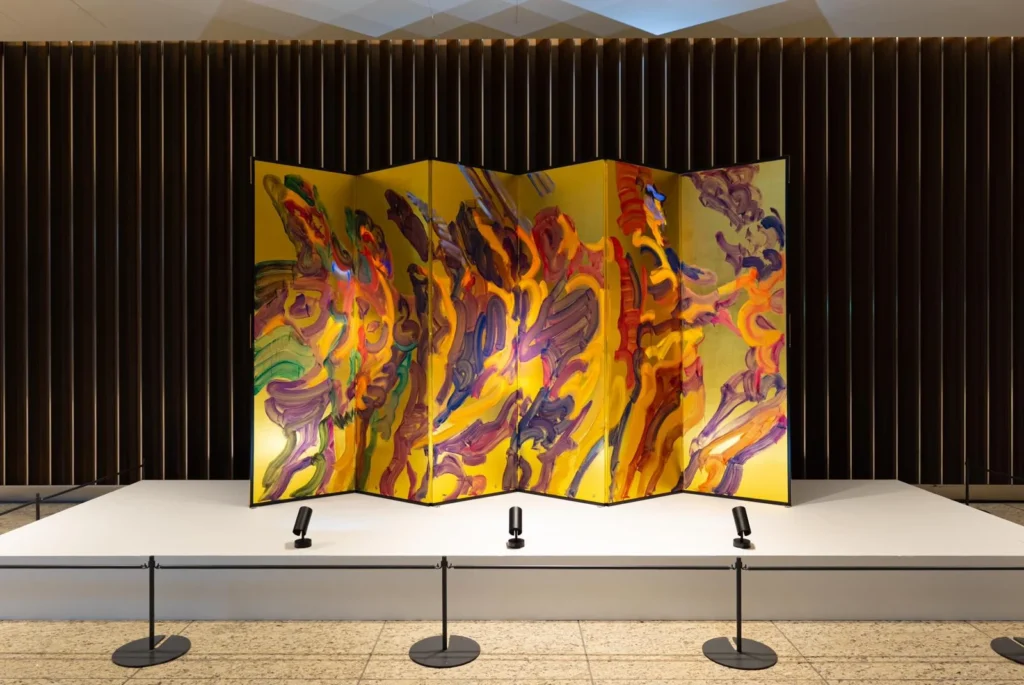 Colorful abstract folding screen displayed on a white platform against a dark slatted wall, with three spotlights in front