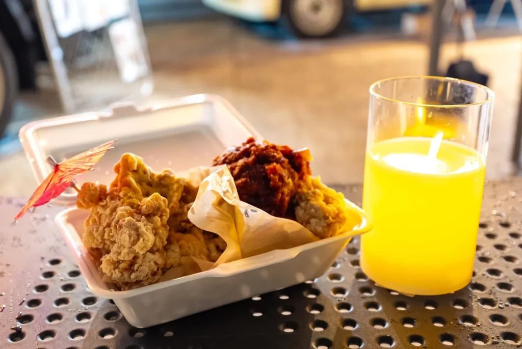 Open takeout box with fried chicken and a bright yellow drink on a perforated metal table