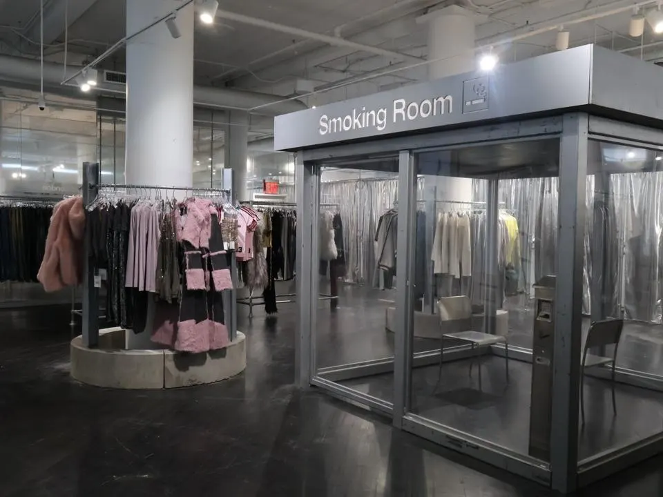 London location clothing racks with pink and black garments beside a glass-enclosed room labeled “Smoking Room” in an industrial showroom
