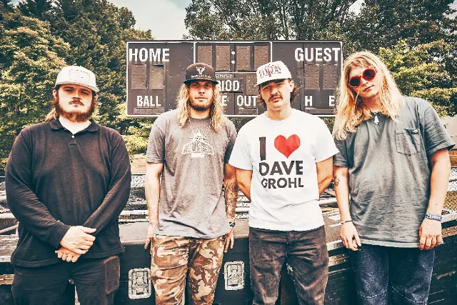 Four musicians pose outdoors before a scoreboard, wearing caps, sunglasses, graphic T-shirts, and casual dark clothing