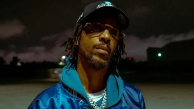 Man with dreadlocks wearing sunglasses, a black cap, blue jacket and layered chains outdoors at dusk