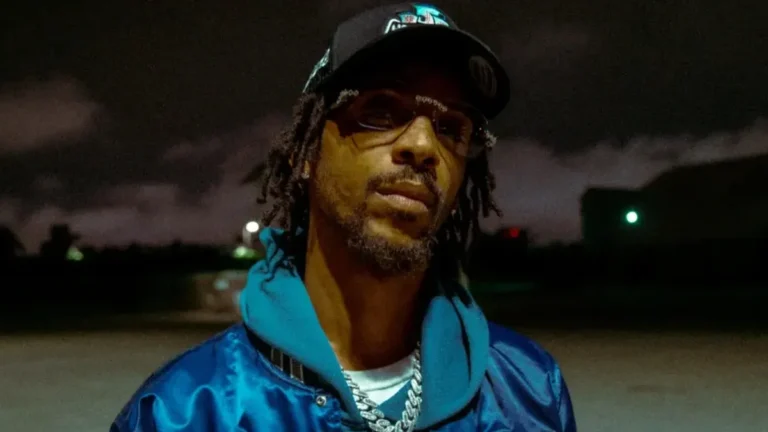 Man with dreadlocks wearing sunglasses, a black cap, blue jacket and layered chains outdoors at dusk