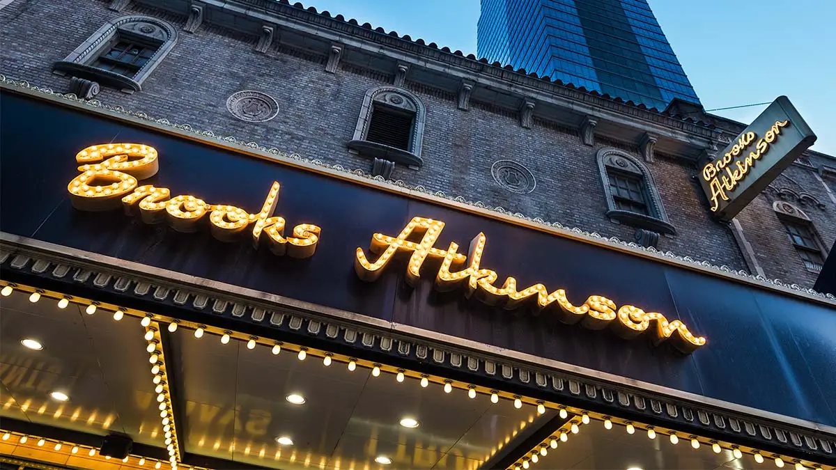 The Brooks Atkinson Theatre facade with glowing marquee lights, ornate brickwork, and a glass skyscraper behind it