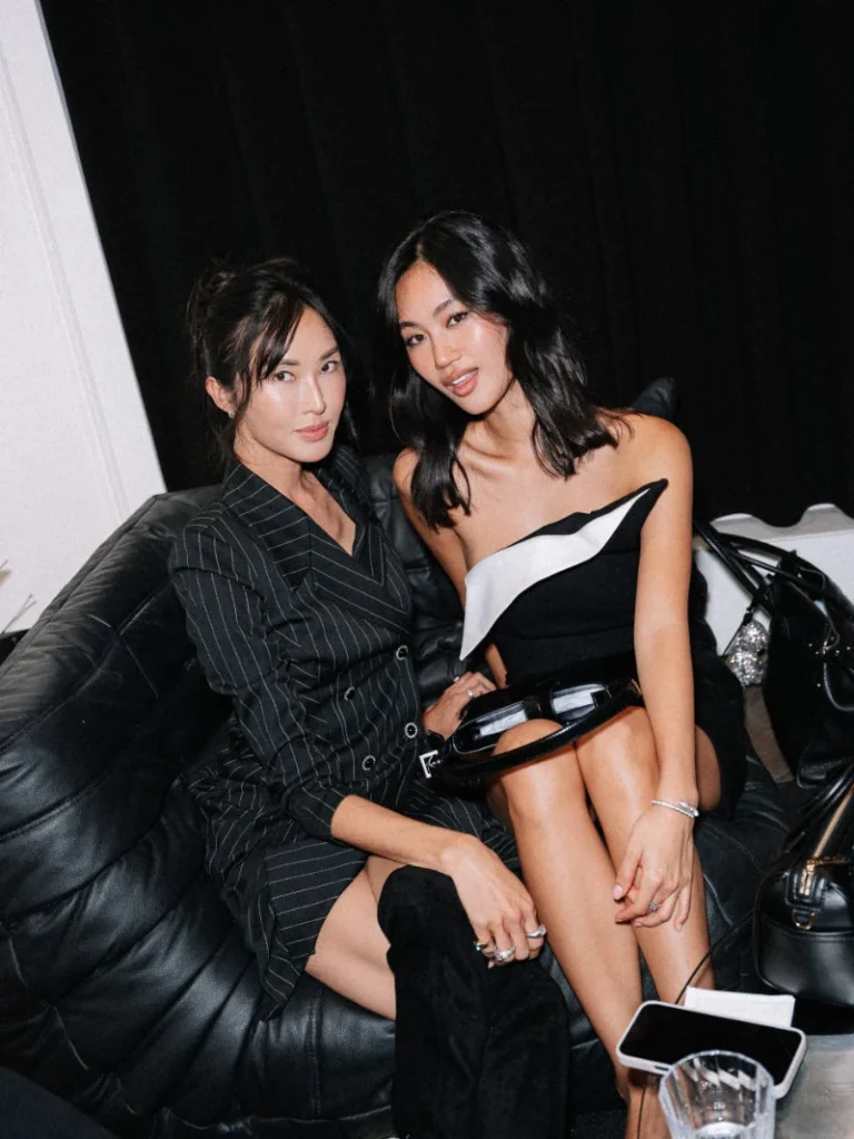 Two women pose on a black leather couch; one wears a pinstriped suit, the other a black-and-white strapless dress