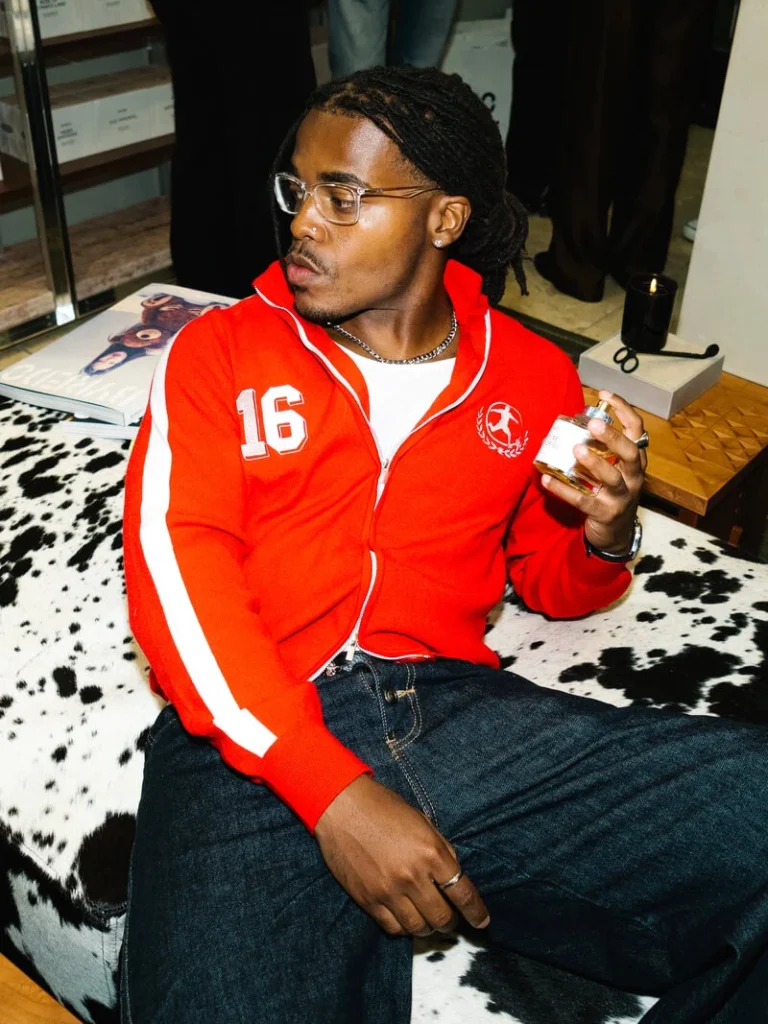 Person with braided hair and glasses sits on a cowhide bench wearing a red zip-up jacket and holding a drink