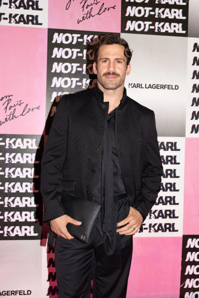 Man in a black textured jacket and shirt holds a black clutch before a pink-and-black Karl Lagerfeld backdrop