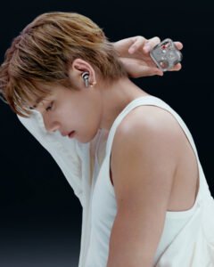 V in a white sleeveless top wearing a black earbud, holding a transparent earbud case against a black background