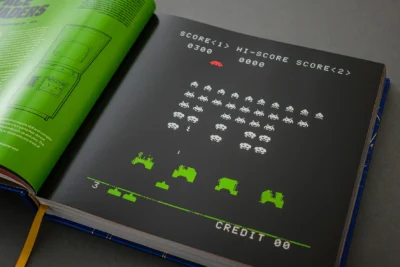 Open book showing a pixel-art arcade game screen with white invaders and green ships on a black page beside a neon-green page