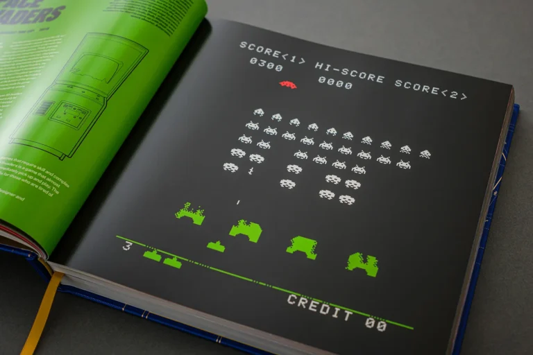 Open book showing a pixel-art arcade game screen with white invaders and green ships on a black page beside a neon-green page