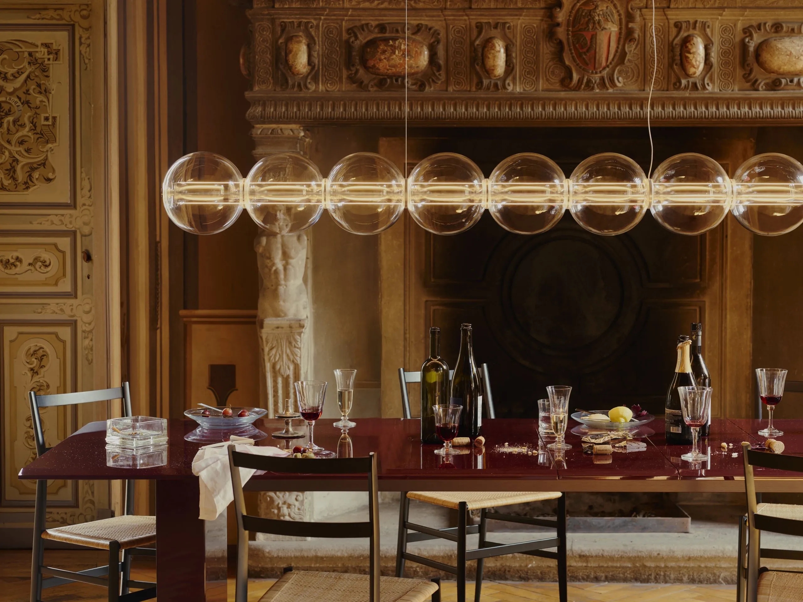 Linear pendant light with clear glass spheres on a glowing horizontal bar above a set dining table in an ornate room