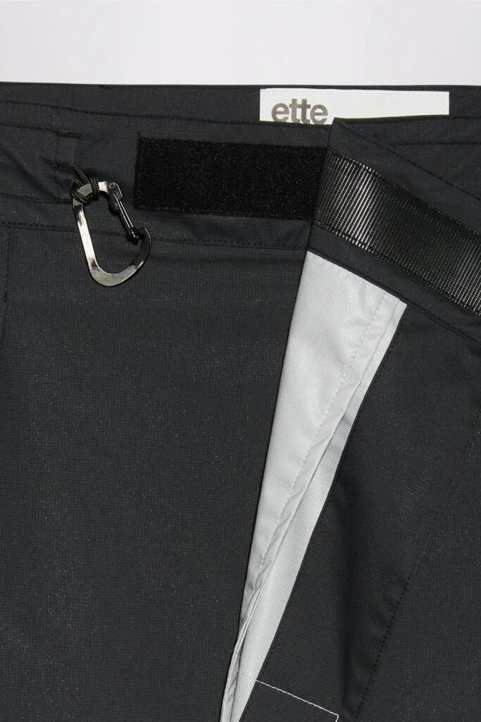 Close-up of black pleated fabric with a carabiner, mesh panel, white insert and ette label