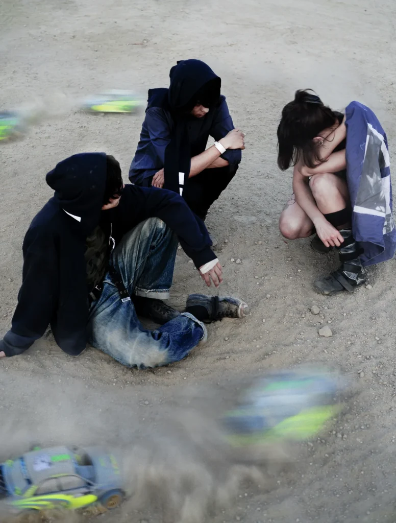 Three people in dark clothing sit on gravel as blurred neon-accented objects pass around them