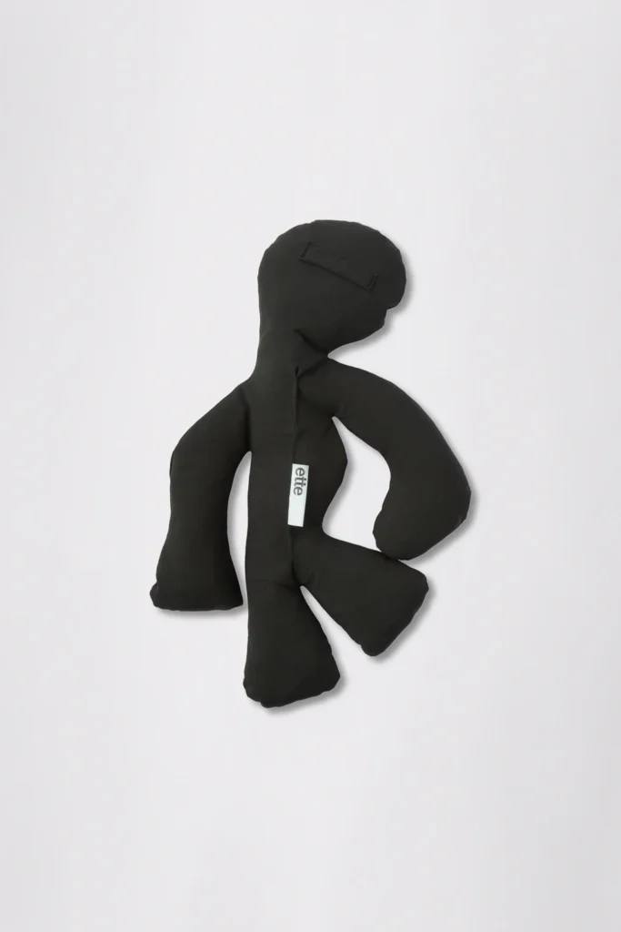 Black fabric doll with stitched features and a white vertical label, posed against a light background