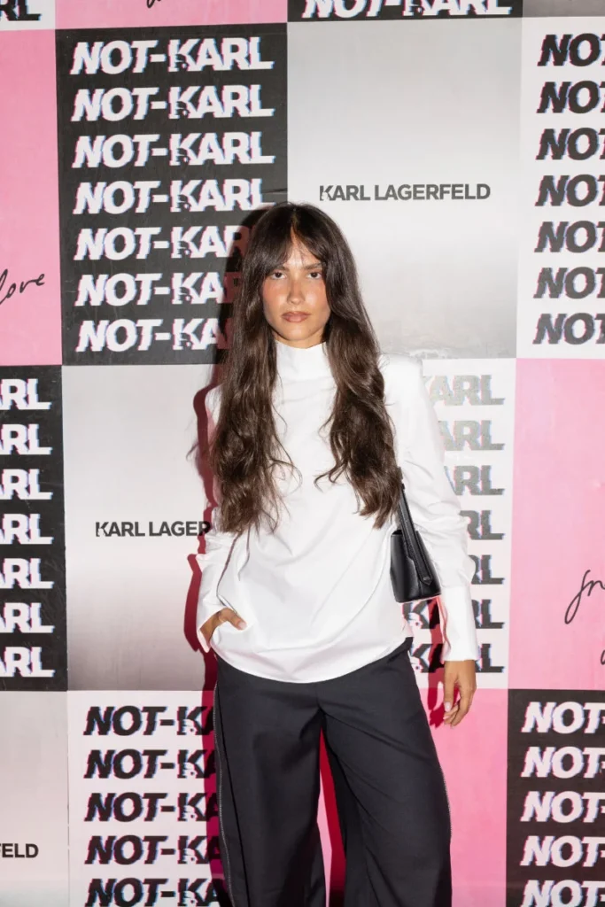 Woman in a white high-neck top and black trousers poses with a black shoulder bag before a pink-and-black Karl Lagerfeld