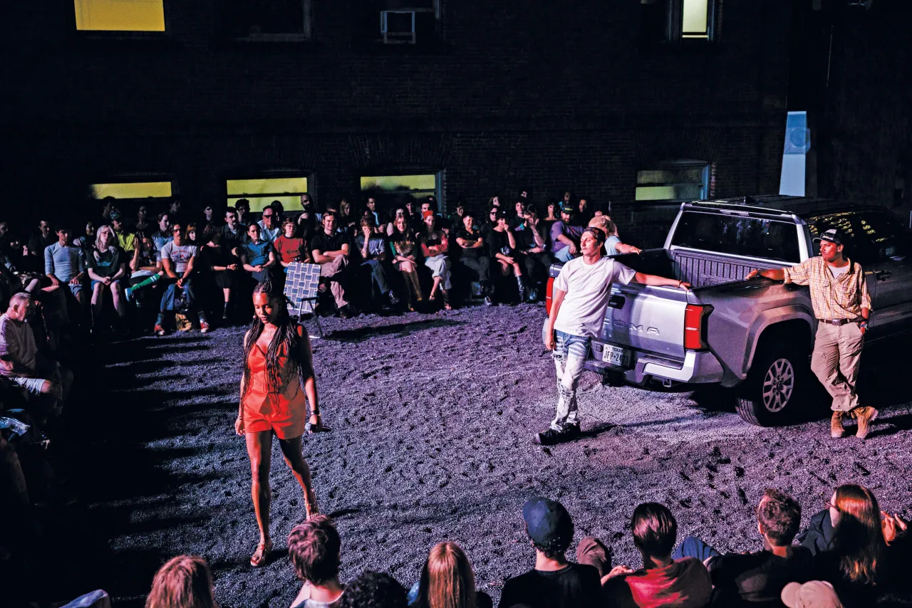 Crowd gathers around a pickup truck in a gravel lot as a woman in orange walks past