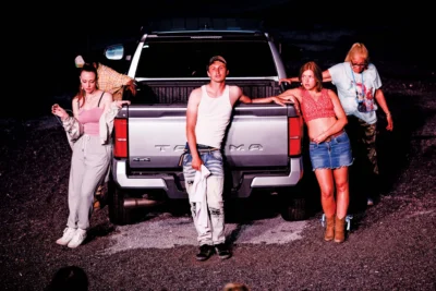 Four people pose around the tailgate of a silver pickup truck at night; one wears a white tank top and ripped jeans