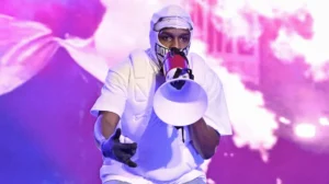 Masked performer in white clothing holds a red-and-white megaphone toward the camera under purple and pink stage lights