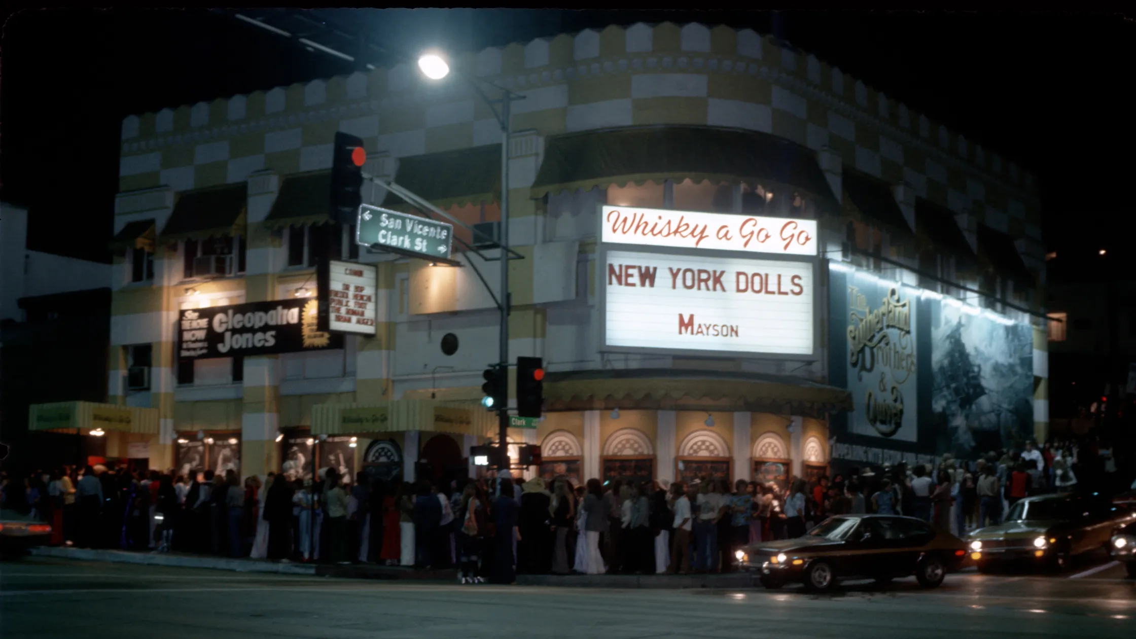 Nighttime street scene outside Whisky a Go Go, with a crowd, cars, traffic lights and signs for New York Dolls and Mayson