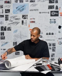 Heron Preston sketches at a table with rolled paper, fabric swatches, tools, and Formula E design boards behind him