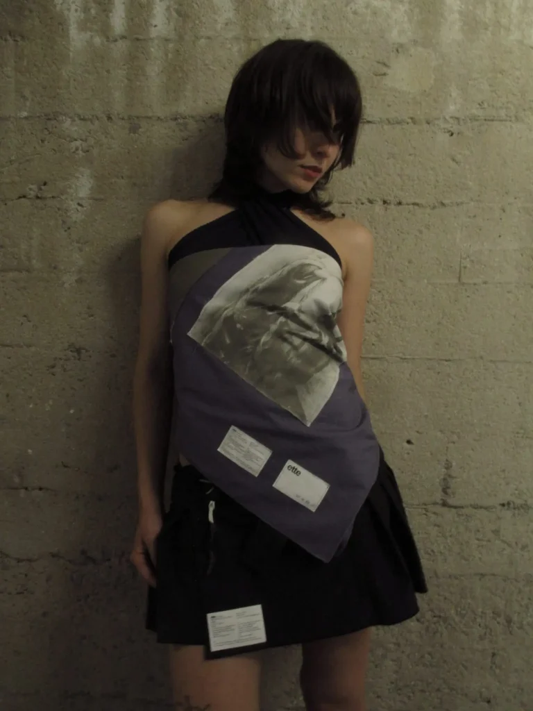 Model with shaggy dark hair wears a purple asymmetrical top with a printed graphic and black skirt against a concrete wall