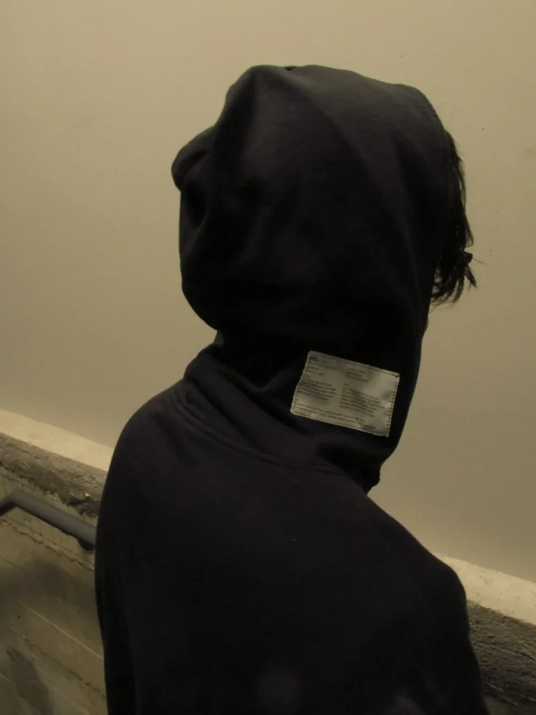 Black hooded figure with face covered and a rectangular label sewn onto the back of the hood