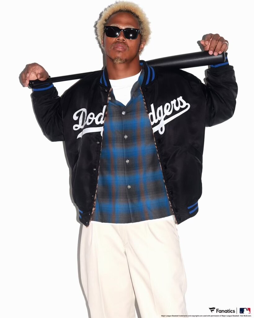Person in sunglasses holds a black bat across his shoulders, wearing a Dodgers varsity jacket, blue plaid shirt and white