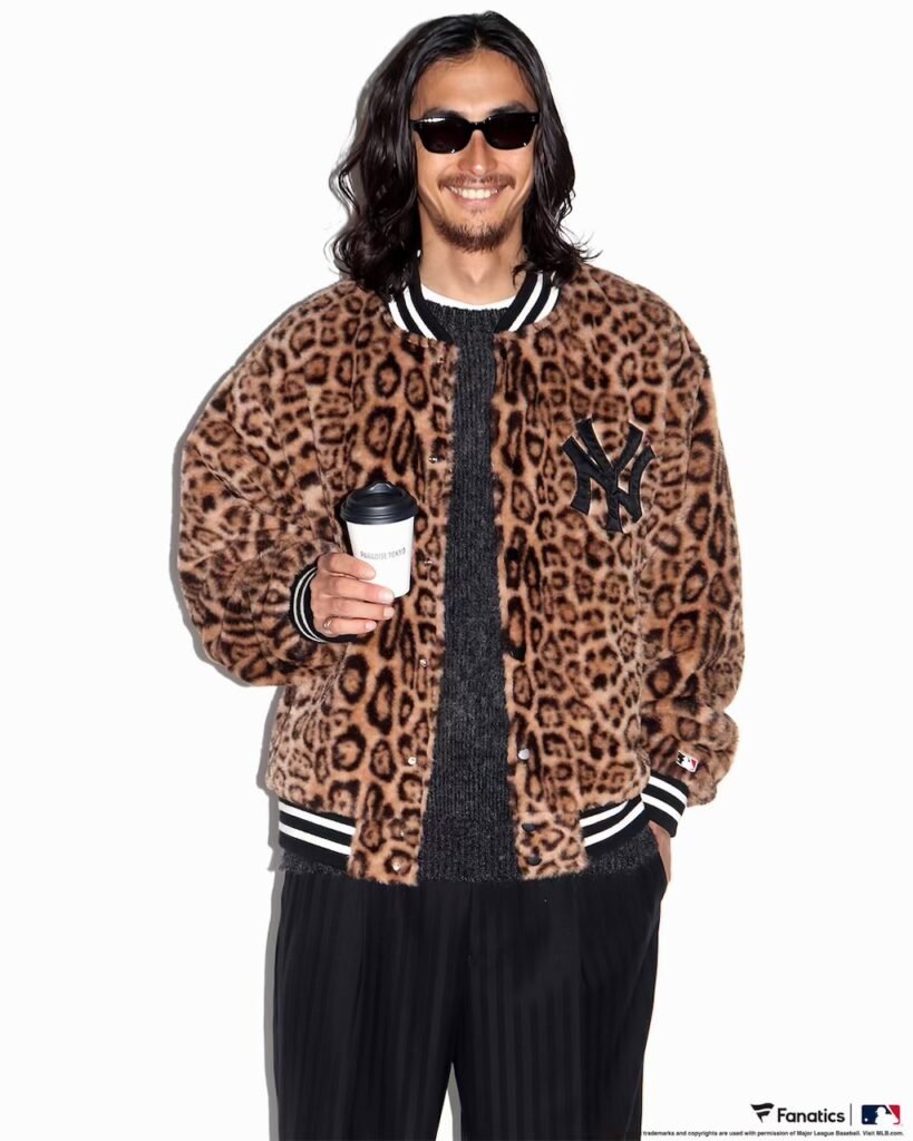 Smiling person in sunglasses wears a brown leopard-print varsity jacket with a New York Yankees patch and holds a coffee cup