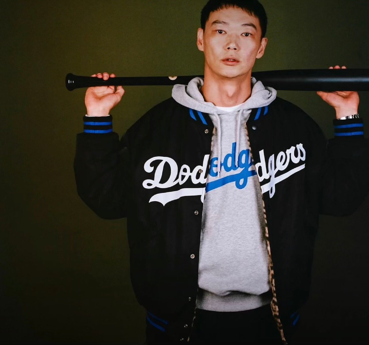 Person holds a black baseball bat across his shoulders, wearing a black Dodgers jacket with leopard-print lining and gray