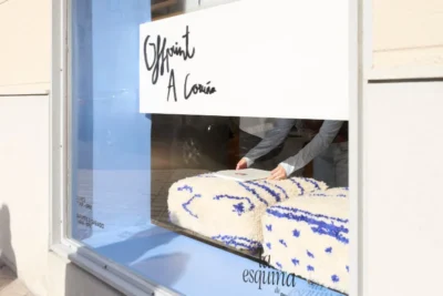 Store window reading “Offprint A Coruña,” with white-and-blue textured textiles displayed and a person arranging them inside