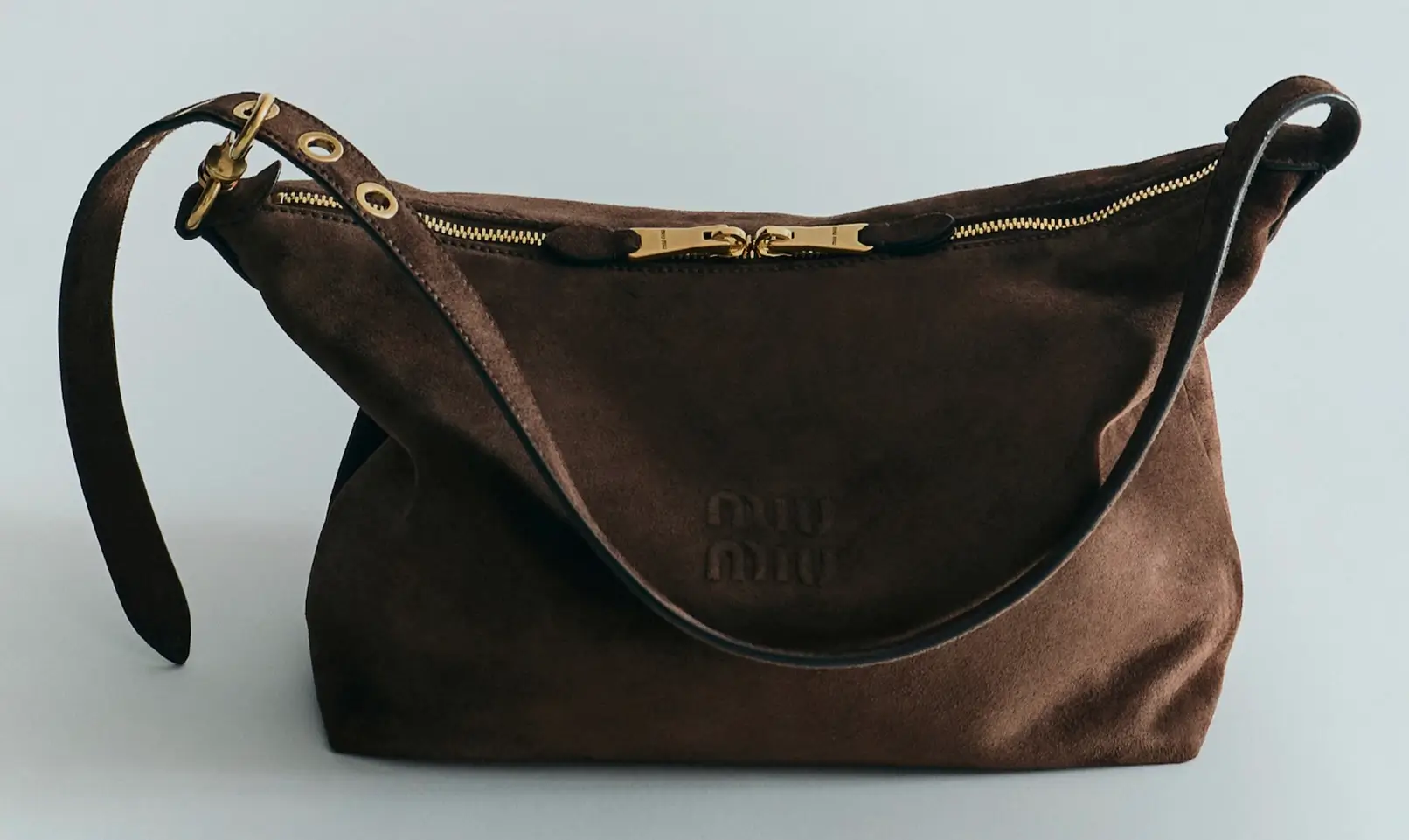 Brown suede shoulder bag with a long strap, gold zipper and hardware, against a pale background