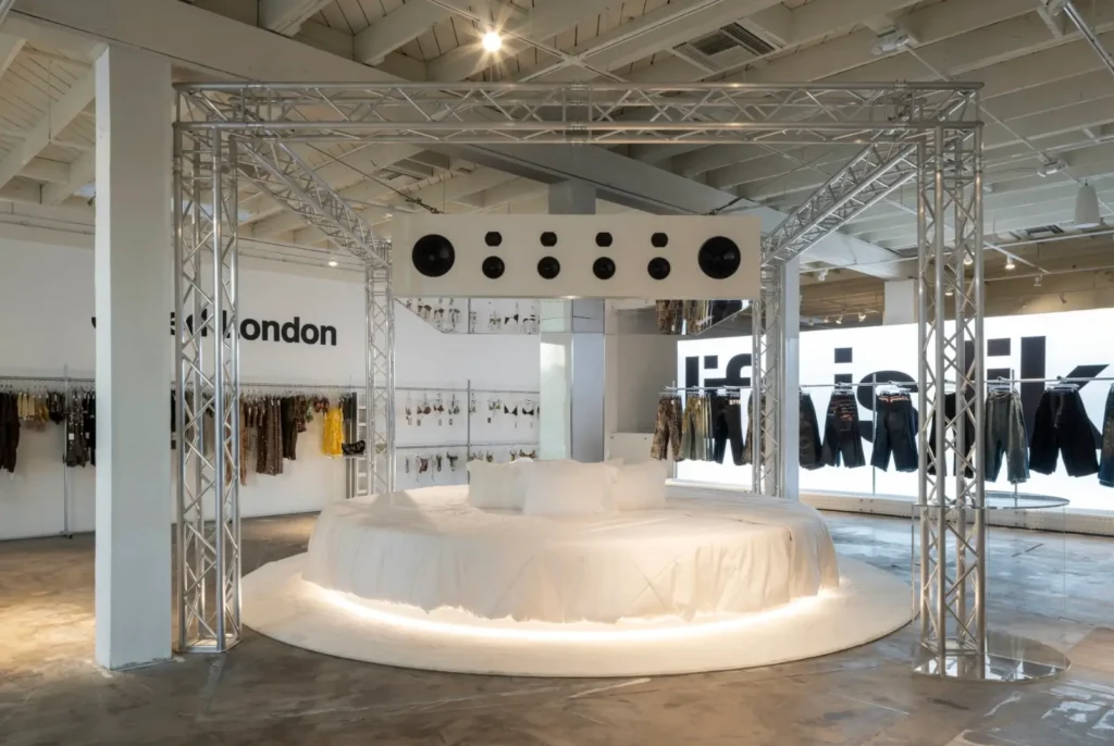 Jaded London showroom with clothing racks, a white circular lounge, and a suspended speaker display framed by metal trusses