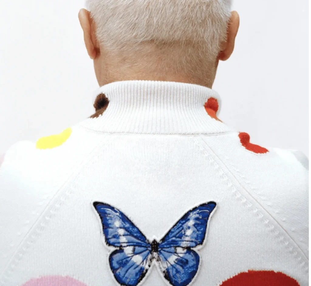 Back view of a white-haired person in a white sweater featuring a large blue butterfly and colorful spots