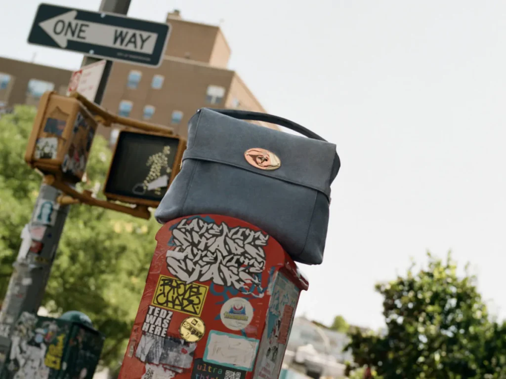 Dark gray handbag with a top handle and gold oval clasp perched on a sticker-covered street box beneath a one-way sign