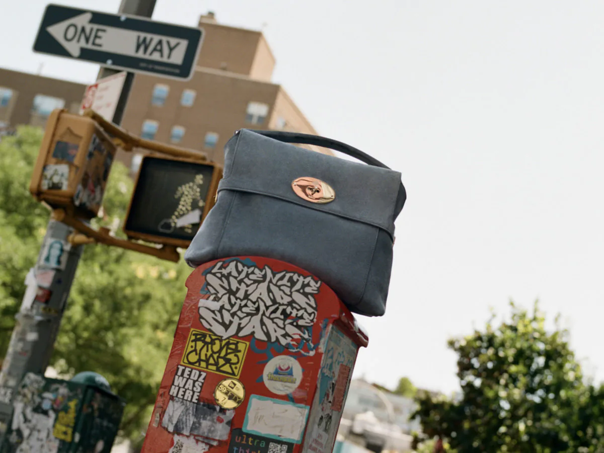Dark gray handbag with a top handle and gold oval clasp perched on a sticker-covered street box beneath a one-way sign