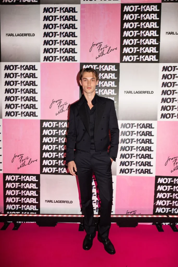 Man in a black suit stands on a pink carpet before a pink-and-black NOT-KARL and Karl Lagerfeld backdrop