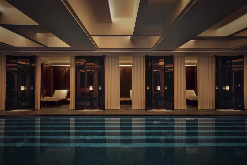 Indoor pool facing curtained treatment rooms with cream loungers, dark wood, mirrored doors, and warm recessed lighting