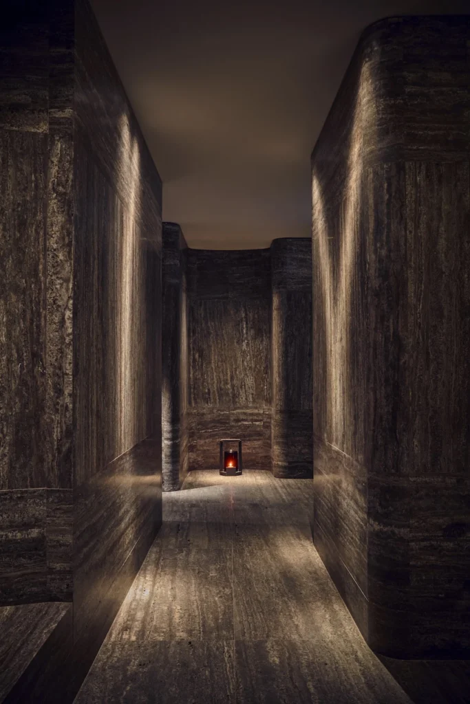 Dimly lit narrow spa corridor with textured dark walls and a small glowing red light at the far end