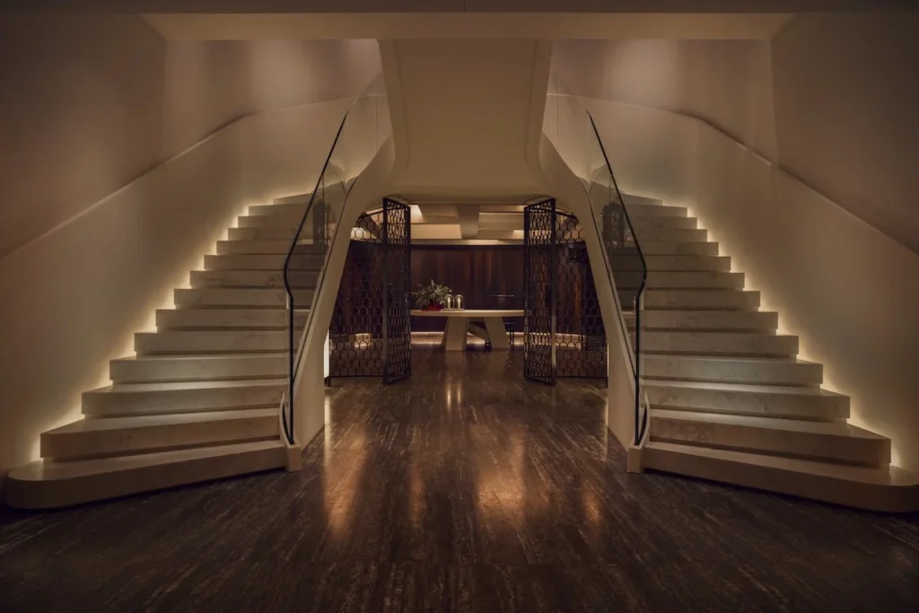 Curved staircases flank a dark wood floor leading to a warmly lit lounge with patterned screens