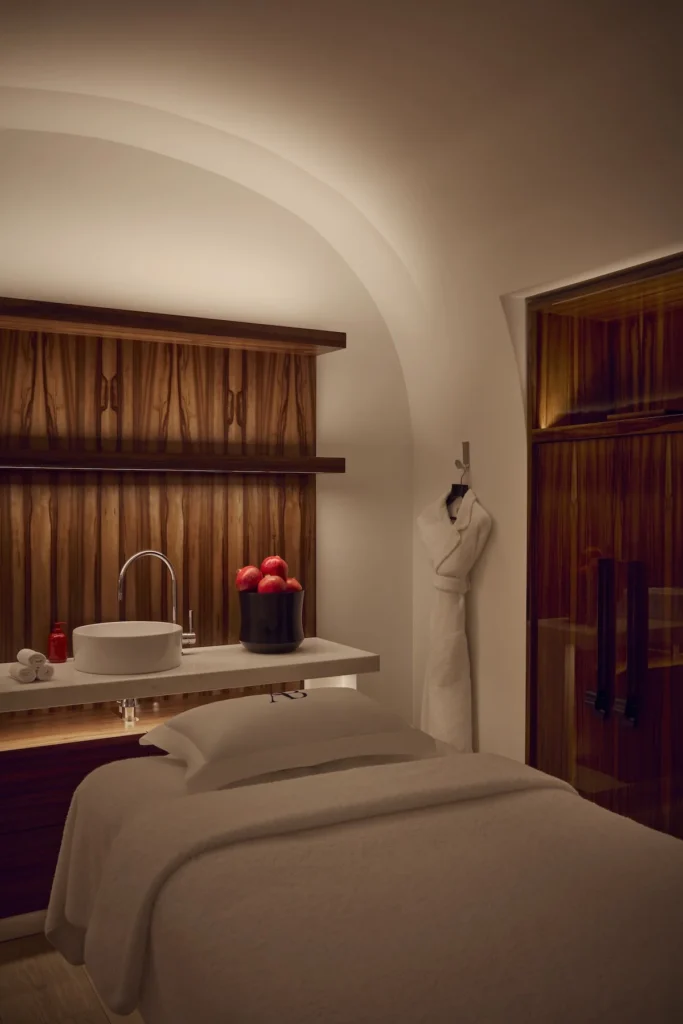 Spa treatment room with a covered bed, rounded plaster ceiling, wooden shelving, sink, and white robe hanging on the wall