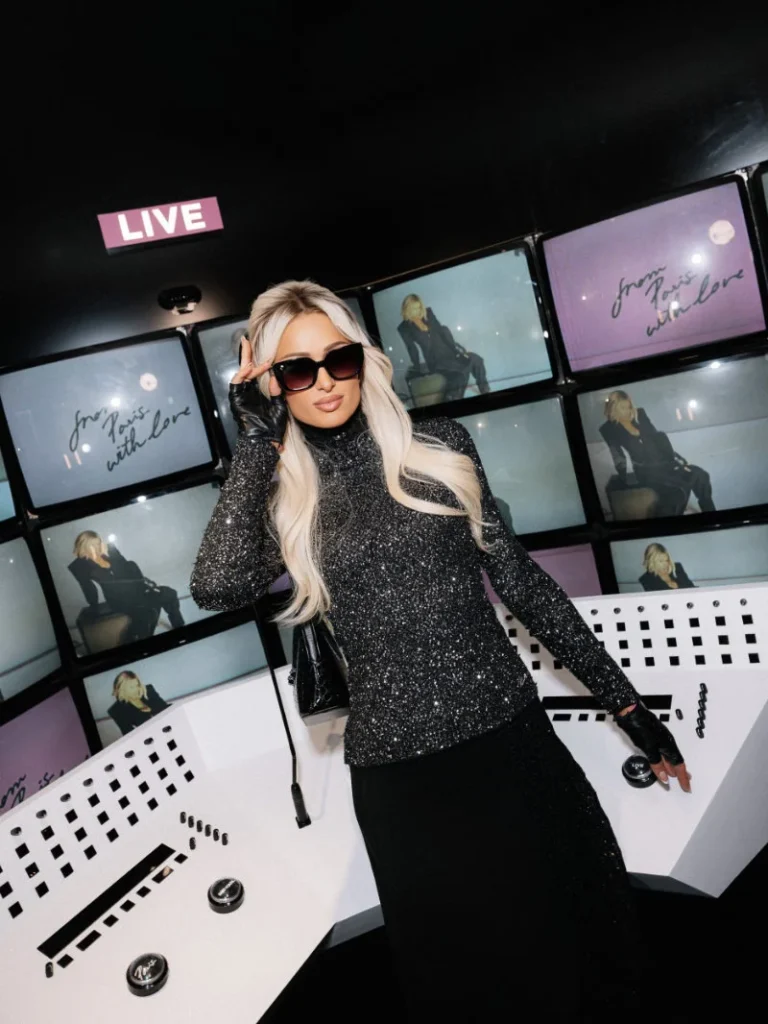 Paris Hilton poses in a glittering black top, long skirt, sunglasses and gloves before screens and a LIVE sign