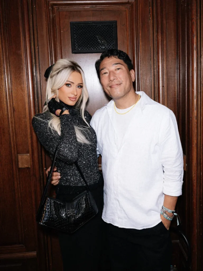 Paris Hilton and Hun Kim pose together before a dark wood-paneled door; Hilton wears a glittering black top and carries a