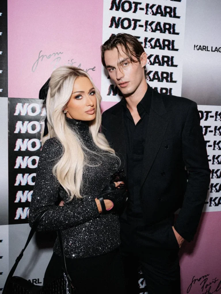 Paris Hilton and a man pose arm-in-arm in black outfits against pink and black NOT-KARL branding walls